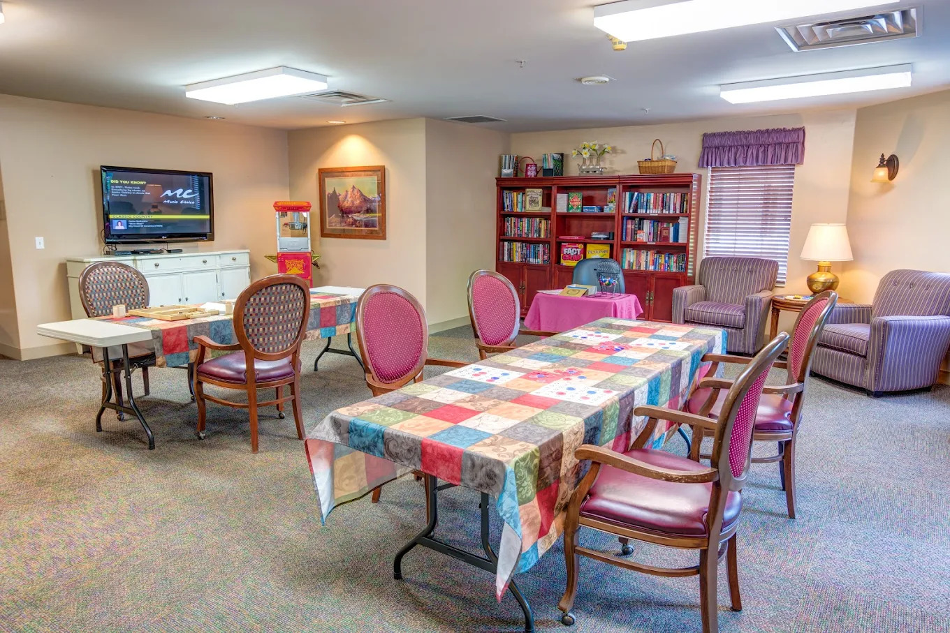 Activity room with a wall television showing a trivia slide, red bookcases of board games, quilt-print tables and a popcorn cart