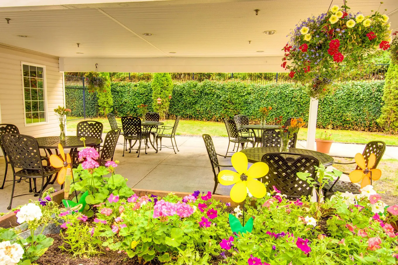 Covered patio with black wrought iron furniture, hanging baskets and a raised bed of petunias and geraniums