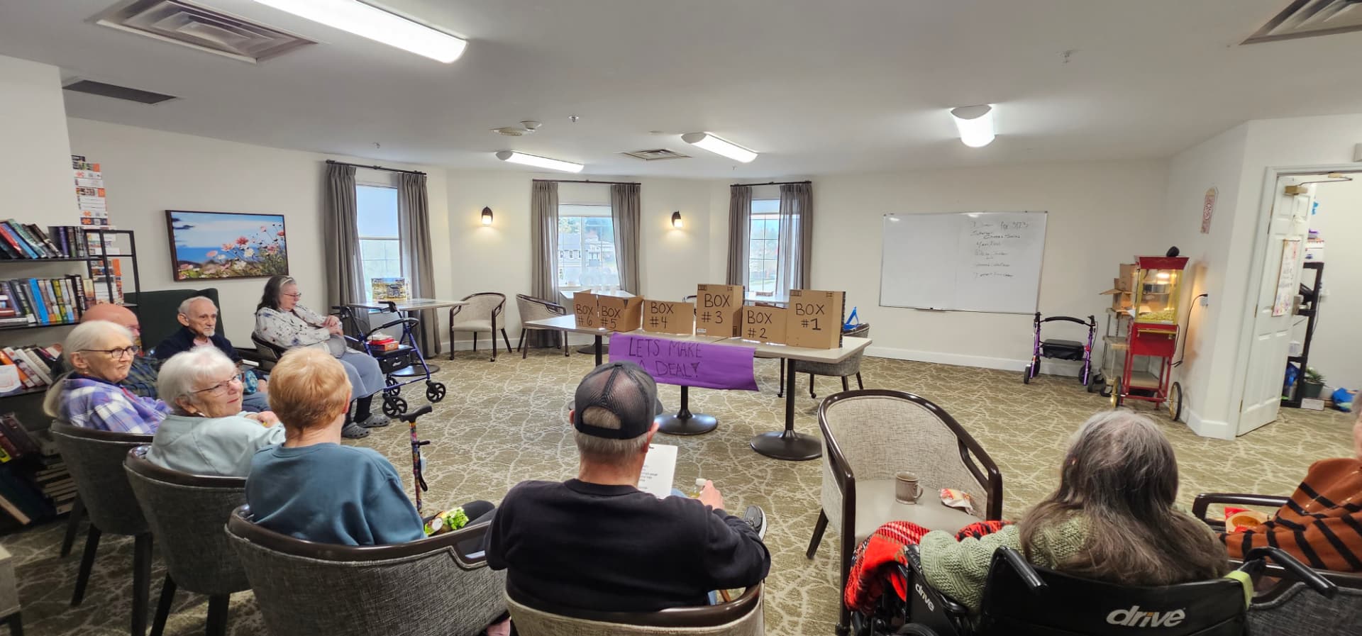 Residents seated in a half circle for a Let's Make a Deal game in the activity room, numbered prize boxes lined up on a table beside the popcorn cart