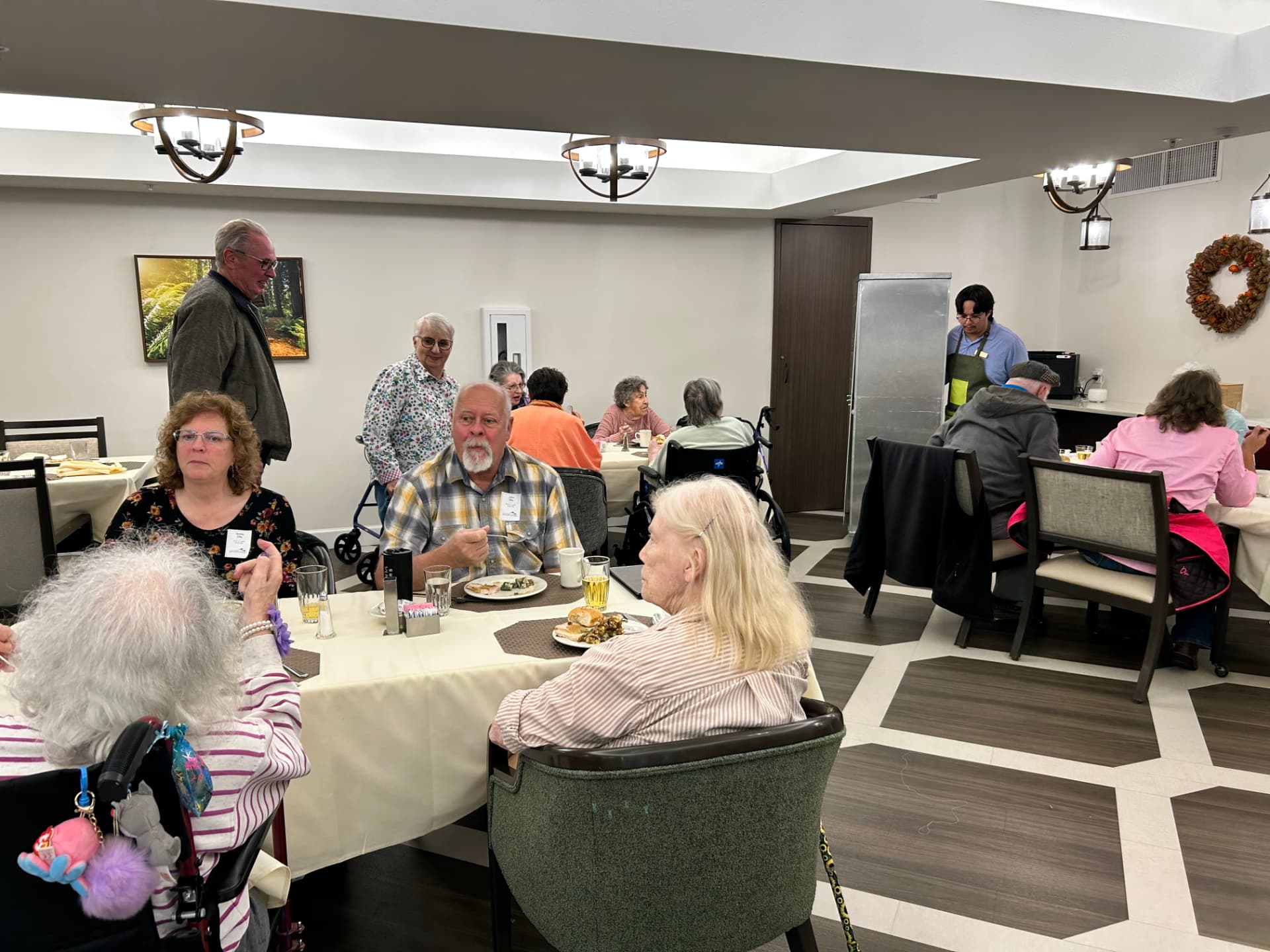 Residents at round tables with white tablecloths during lunch service in the main dining room, chandeliers overhead and staff serving