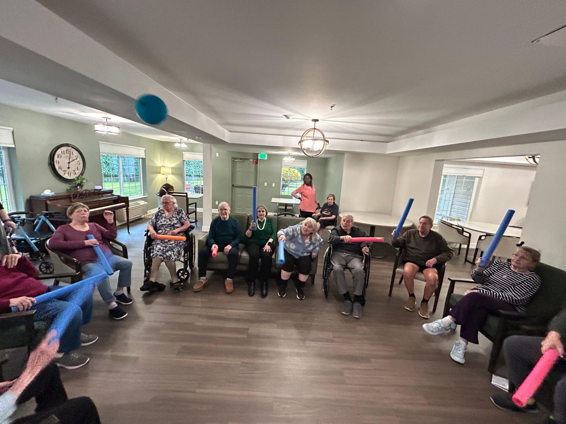 Residents seated in a circle lifting colourful pool noodles during an exercise class in the lounge, a piano and wall clock behind them