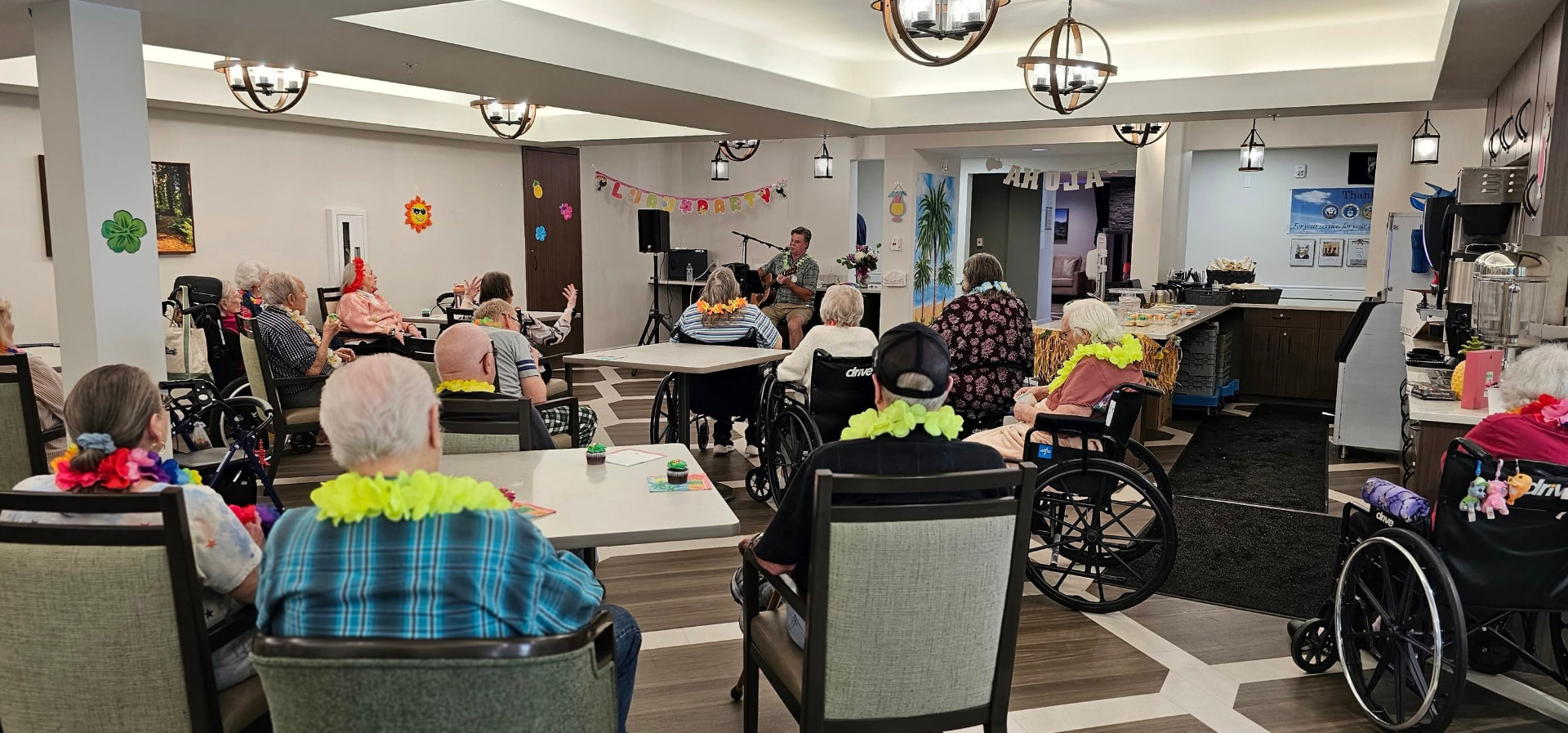 Residents of Cascade Valley Senior Living in Arlington, Washington, wearing flower leis and seated in chairs and wheelchairs in the activity room while a guitarist plays at a luau party beneath a Luau Party banner